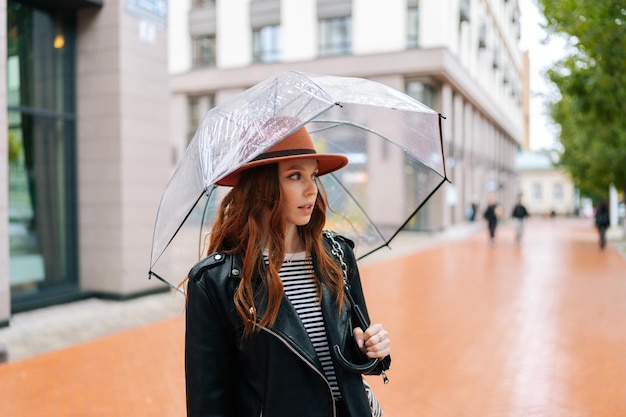 Portrait d'une jolie jeune femme rousse portant un chapeau de mode debout avec un parapluie transparent sur la rue de la ville près du centre par temps de pluie pluie