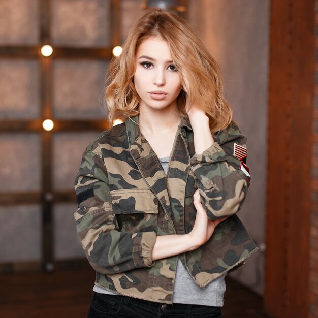 Portrait d'une jolie jeune femme sexy avec de beaux yeux dans une veste de camouflage à la mode et un t-shirt gris dans le studio sur le fond d'un mur en bois et de lumières. Blonde élégante et bouclée