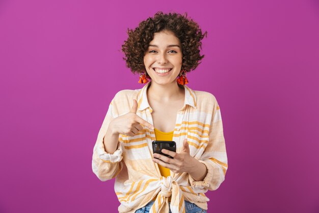 Portrait d'une jolie jeune femme souriante aux cheveux bouclés brune debout isolée sur un mur violet, pointant sur un téléphone portable