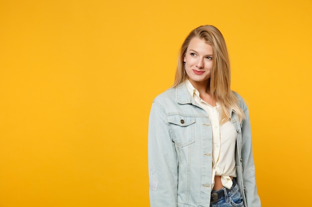 Portrait d'une jolie jeune femme souriante dans des vêtements décontractés en denim debout et regardant de côté isolé sur fond de mur orange jaune vif en studio. Concept de style de vie des gens. Maquette de l'espace de copie.