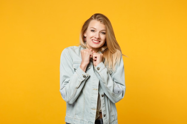 Portrait d'une jolie jeune femme souriante dans des vêtements décontractés en denim regardant la caméra, serrant les poings isolés sur un mur de fond orange jaune vif en studio. Concept de style de vie des gens. Maquette de l'espace de copie.