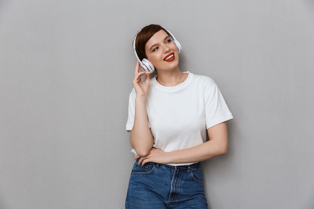 Portrait d'une jolie jeune femme souriante en écoutant de la musique avec des écouteurs isolés sur un mur gris