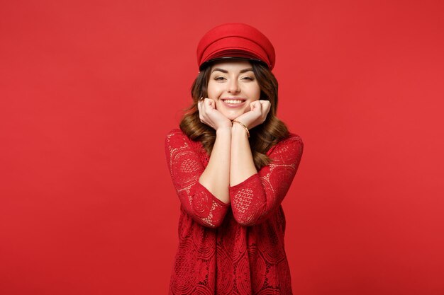 Portrait d'une jolie jeune femme souriante en robe de dentelle et bonnet mis la main sur le menton isolé sur fond de mur rouge vif en studio. Émotions sincères des gens, concept de style de vie. Maquette de l'espace de copie.