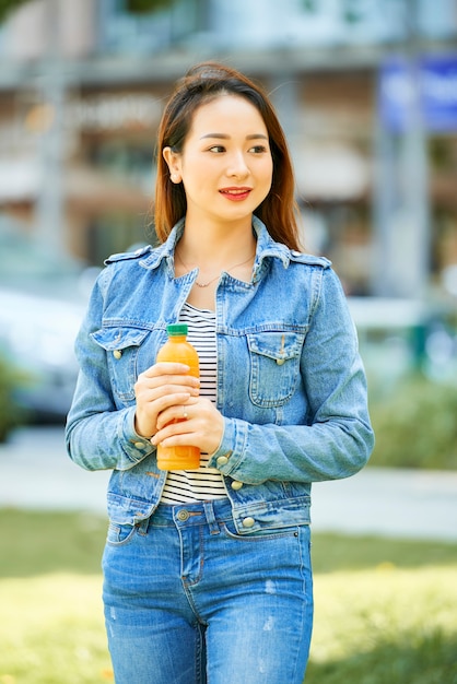 Portrait d'une jolie jeune femme souriante veste en jean debout à l'extérieur et ouvrant une bouteille en plastique de ...