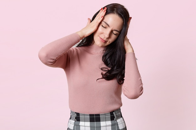 Portrait de jolie jeune femme avec un terrible mal de tête, garde les deux mains sur la tête, doit prendre des médicaments, femme brune en chemise rose et jupe à carreaux, isolé sur rose