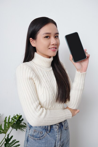 Portrait d'une jolie jeune femme vietnamienne en pull blanc montrant un smartphone avec un écran vide vierge