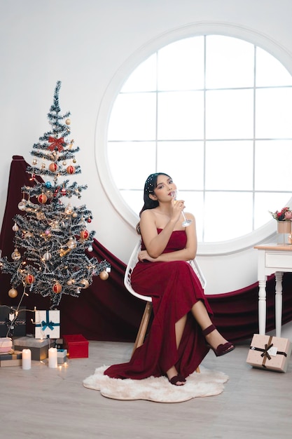 Portrait de jolie jeune fille confortable s'asseoir et boire du vin porter une robe rouge dans le salon de Noël décoré à l'intérieur