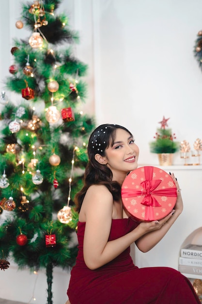 Portrait de jolie jeune fille confortable s'asseoir tenant le cadeau de Noël souriant porter une robe rouge dans le salon de Noël décoré à l'intérieur