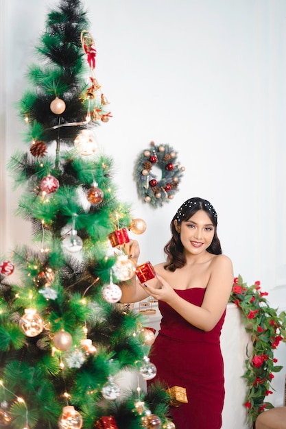 Portrait d'une jolie jeune fille décorant un sapin de Noël souriante vêtue d'une robe rouge dans un salon de Noël décoré à l'intérieur