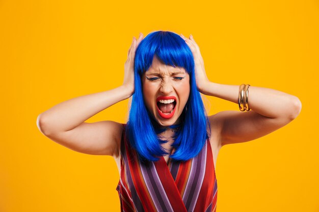 Portrait d'une jolie jeune fille funky agacée aux cheveux bleus portant une tenue décontractée isolée sur un mur jaune, couvrant les oreilles, criant