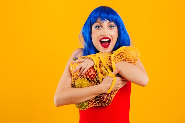 Portrait d'une jolie jeune fille funky excitée aux cheveux bleus portant un maillot de bain isolé sur un mur jaune, sac de transport avec des fruits