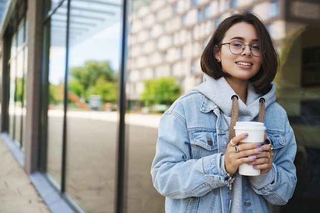 Portrait d'une jolie jeune fille queer en veste en jean marchant au printemps à la recherche d'un appareil photo heureux avec un sourire heureux mur de verre de construction maigre et boire du café