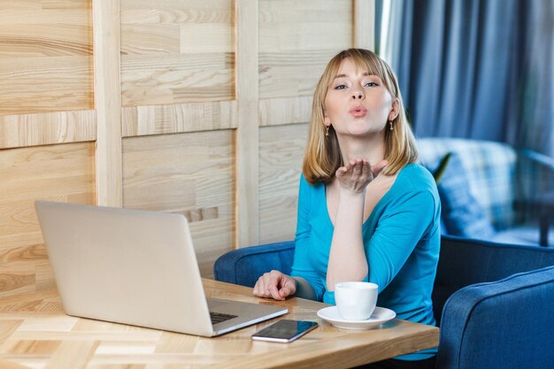 Portrait d'une jolie jeune fille romantique indépendante avec des cheveux blonds coupés en chemisier bleu sont assis seuls dans un café et travaillent sur un ordinateur portable et envoient un baiser aérien, regardant la caméra.