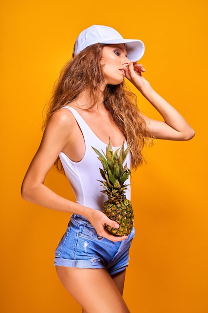 Portrait d'une jolie jeune fille en tenue de plage tenant ananas isolé sur fond jaune.