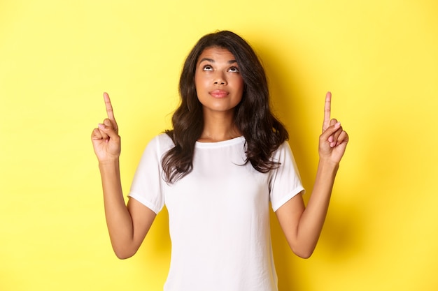 Portrait d'une jolie mannequin afro-américaine, vêtue d'un t-shirt blanc, regardant et pointant les doigts vers le haut avec un sourire curieux, montrant une promo, debout sur fond jaune.