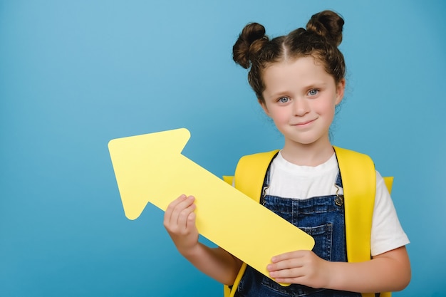 Portrait d'une jolie petite écolière tenant une flèche jaune pointant sur l'espace de copie pour le contenu promotionnel, porte un sac à dos, posant isolé sur un mur de fond bleu studio. Retour au concept de l'école