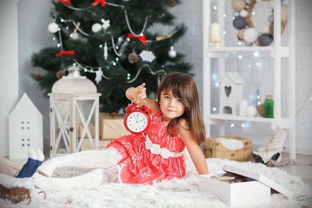 Portrait d'une jolie petite fille brune tenant un réveil rouge dans les mains à l'intérieur avec des décorations de Noël