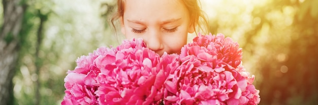 Portrait d'une jolie petite fille caucasienne mignonne de sept ans, tient dans les mains et sent et profite d'un bouquet de fleurs de pivoine rose en pleine floraison sur fond de nature. bannière. éclater