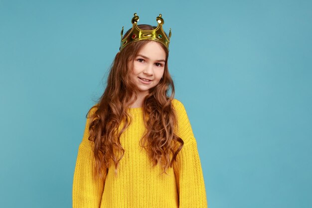 Portrait de jolie petite fille en couronne de diadème doré debout regardant la caméra avec un sourire à pleines dents portant un chandail de style décontracté jaune tourné en studio intérieur isolé sur fond bleu