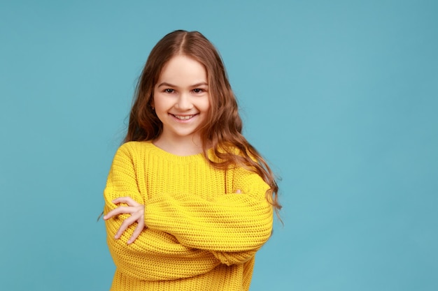 Portrait d'une jolie petite fille debout avec les bras croisés et regardant la caméra avec une expression heureuse, portant un pull jaune de style décontracté. Studio intérieur tourné isolé sur fond bleu.