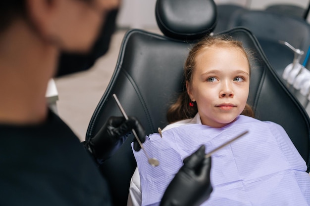 Portrait d'une jolie petite fille effrayée assise dans un fauteuil dentaire et regardant avec peur une dentiste méconnaissable tenant dans les mains des instruments dentaires