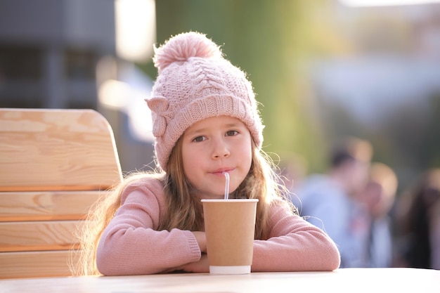 Portrait d'une jolie petite fille enfant au chapeau rose assise seule au café de la rue buvant du thé dans une tasse en papier