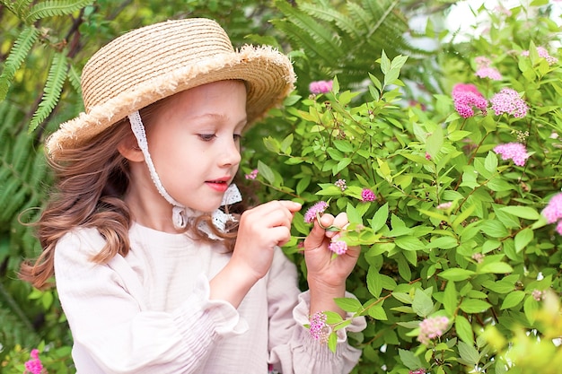 Portrait d'une jolie petite fille européenne vêtue d'une robe beige, chapeau de paille un jour d'été dans un parc de la ville près de parterres de fleurs aux couleurs vives. Moments lumineux de l'enfance.