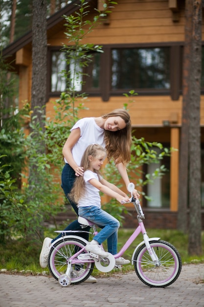 Portrait de jolie petite fille fille souriante et mère vélo vélo dans une cour journée d'été ensoleillée. Loisirs actifs en famille avec des enfants.