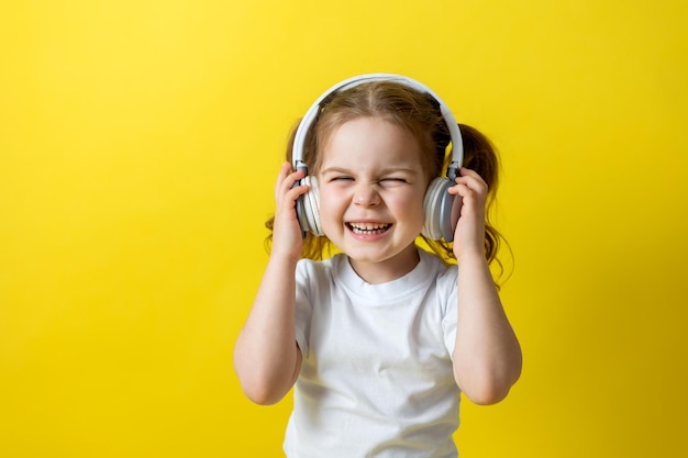 Portrait d'une jolie petite fille gaie écoutant de la musique avec un casque blanc. livres audio, leçons audio. notion d'éducation. studio photo, fond jaune, espace de texte. Photo de haute qualité