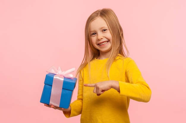 Portrait d'une jolie petite fille ravie et heureuse pointant vers une boîte-cadeau offrant un cadeau d'anniversaire et souriant joyeusement à la caméra son rêve d'enfant devenu réalité studio d'intérieur isolé sur fond rose