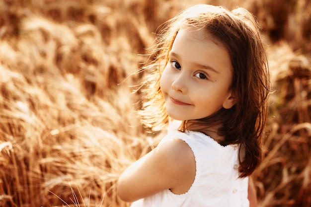 Portrait d'une jolie petite fille vêtue d'une robe blanche touchant le blé contre le coucher du soleil en souriant.