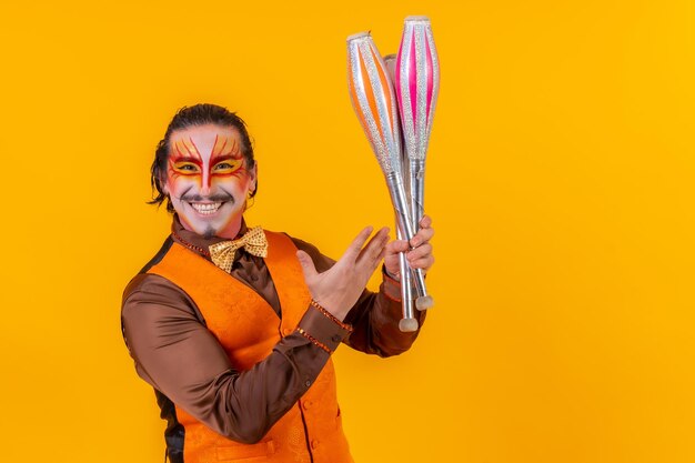 Portrait d'un jongleur dans un gilet avec un visage peint jonglant avec des masses sur fond jaune