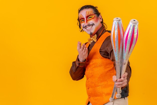 Portrait d'un jongleur dans un gilet avec un visage peint jonglant avec des masses sur fond jaune