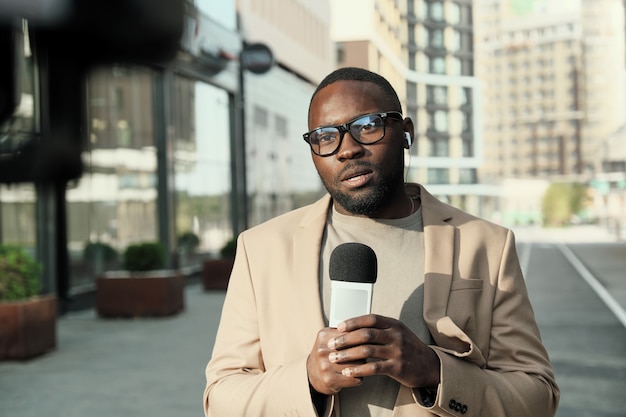 Portrait de journaliste africain à lunettes parlant au microphone tout en travaillant dans la ville