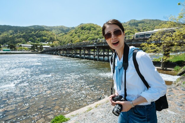 portrait joyeuse femme asiatique photographe sourit à la caméra près du pont Togetsu-kyo à Arashiyama, au japon avec une eau de rivière cristalline à l'arrière-plan.