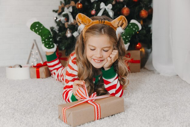 Portrait d'une joyeuse fille de 9 ans, allongée sur le sol près de boîtes avec des cadeaux et un sapin de Noël à la maison. une fille dans une jante avec des oreilles de tigre.