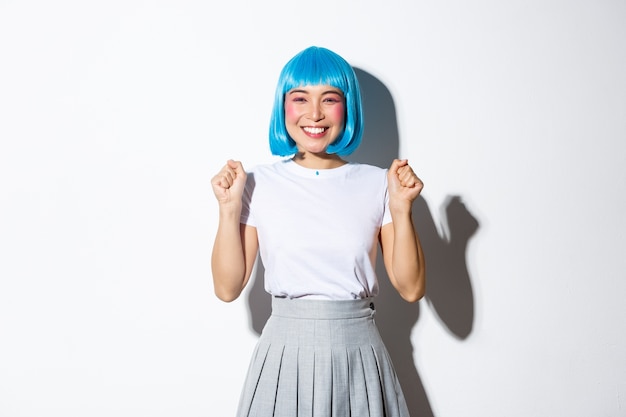 Portrait de joyeuse fille asiatique kawaii en perruque de fête bleue célébrant la victoire, souriant heureux et sautant de se réjouir, triomphant du succès, debout.
