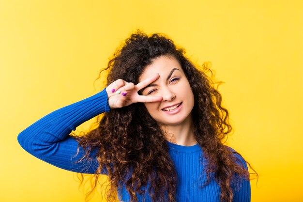 Portrait de joyeuse fille brune aux cheveux bouclés. Dame émotionnelle drôle cligne des yeux à travers les doigts dans le geste de la victoire, souriant.