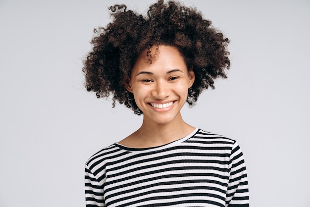 Portrait de joyeuse fille frisée multiraciale dans des vêtements décontractés souriant sincèrement à la caméra. Concept d'humeur positive. Prise de vue en studio intérieur, isolé sur fond blanc