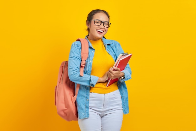 Portrait de joyeuse jeune étudiante asiatique en vêtements décontractés avec sac à dos tenant un livre et regardant la caméra isolée sur fond jaune