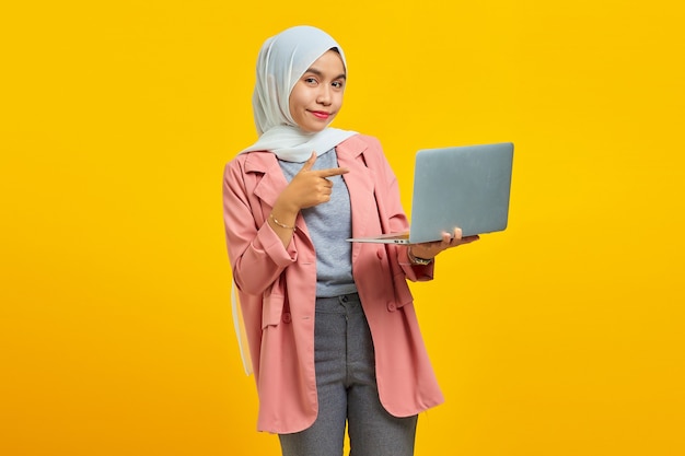 Portrait de joyeuse jeune femme asiatique tenant un ordinateur portable et pointant isolé sur fond jaune