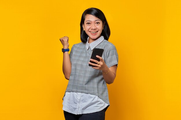 Portrait de joyeuse jeune femme asiatique tenant un téléphone portable et célébrant le succès sur fond jaune