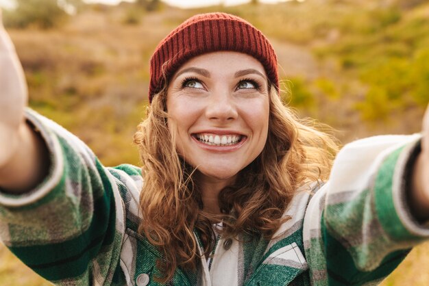 Photo portrait d'une joyeuse jeune femme caucasienne portant un chapeau et une chemise à carreaux prenant un selfie portrait en marchant à l'extérieur