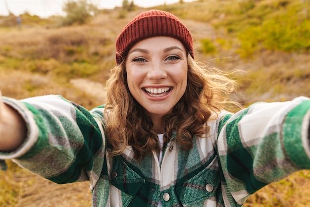 Portrait d'une joyeuse jeune femme caucasienne portant un chapeau et une chemise à carreaux prenant un selfie Portrait en marchant à l'extérieur