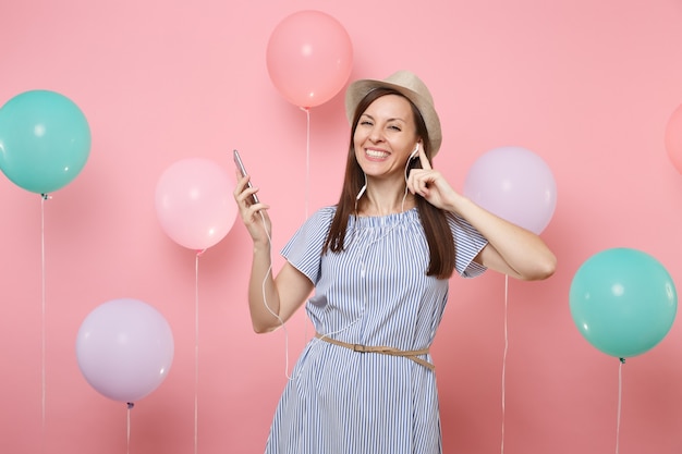 Portrait de joyeuse jeune femme heureuse en chapeau d'été de paille et robe bleue avec téléphone portable et écouteurs écoutant de la musique sur fond rose avec des ballons à air colorés. Concept de fête d'anniversaire.