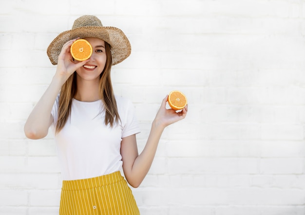 Portrait d'une joyeuse jeune fille au chapeau tenant deux tranches d'orange sur un mur blanc