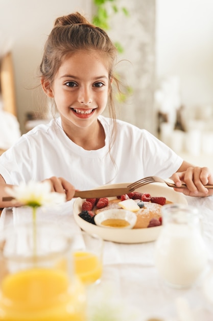 Portrait de joyeuse petite fille assise à table dans une cuisine lumineuse tout en prenant son petit-déjeuner et en mangeant des crêpes à la framboise