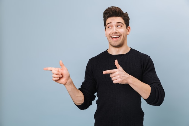 Portrait d'un joyeux beau jeune homme debout isolé sur bleu, pointant vers l'espace de copie