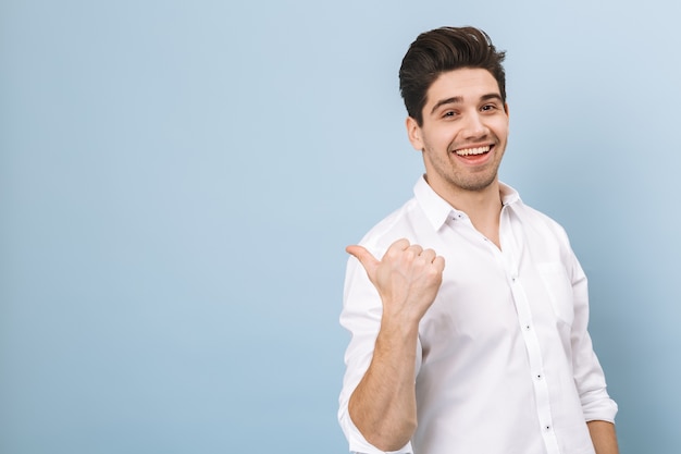 Portrait d'un joyeux beau jeune homme debout isolé sur bleu, pointant vers l'espace de copie