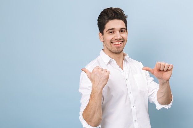 Portrait d'un joyeux beau jeune homme debout isolé sur bleu, pointant vers l'espace de copie